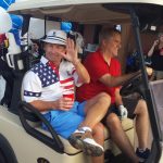 Two people on a golf cart, passenger waving at the camera.