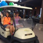 Two people on a golf cart waving to a crowd of camera